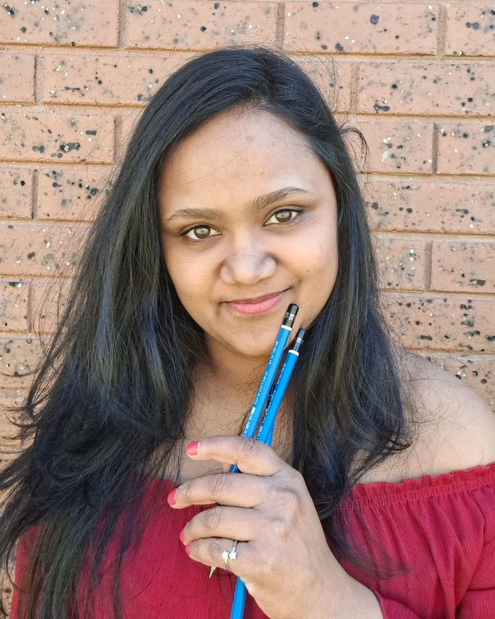 Shamalka, smiling and holding two pencils, in front of a brick wall